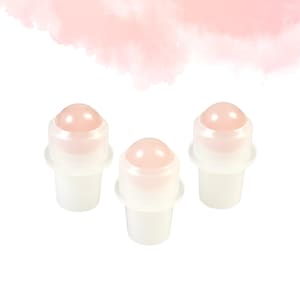 May include: Three roll-on bottles with pale pink gemstone tops. The bottles have a white base and a clear, rounded top. The background features a soft, pink watercolour wash.
