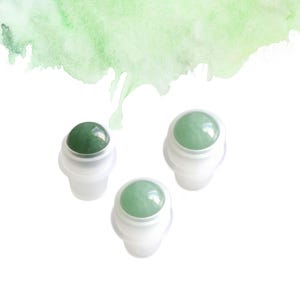May include: Three jade roller bottles with white plastic bases are arranged on a white background. Each bottle has a smooth, round, green jade stone at the top. A green watercolour wash is in the upper left corner.