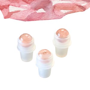 May include: Three rose quartz roller bottles with white plastic components. The rose quartz spheres are a soft pink colour. The bottles are set against a white background, with a watercolour wash in shades of pink at the top.