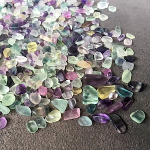 May include: A collection of small, colorful, tumbled fluorite stones in shades of green, purple, and yellow. The stones are scattered on a gray surface.