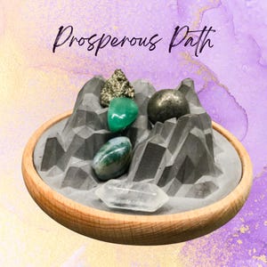 May include: A wooden-rimmed tray holds a gray mountain-shaped structure with various gemstones. The gemstones include a green stone, a clear crystal, and a gold-flecked stone. The text "Prosperous Path" is written in cursive above the tray.