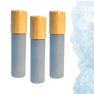 May include: Three cylindrical bottles with light gray bodies and natural wood caps. The bottles are arranged vertically against a white background with a blue watercolor wash on the right side. The wood caps have a visible grain.