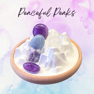 May include: A wooden tray holds a white mountain-shaped sculpture and several polished gemstones in shades of purple, blue, and clear. The text "Peaceful Peaks" is written in cursive above the tray.