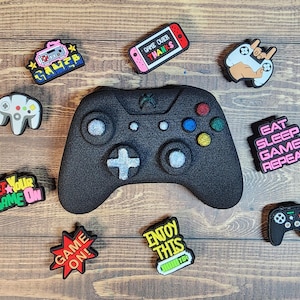 May include: A black Xbox controller with various colorful game-themed shoe charms. The charms include phrases like "Game On!" and "Eat Sleep Game Repeat".
