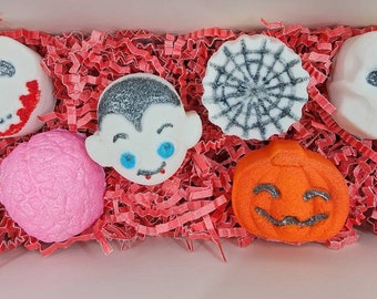 May include: A box filled with Halloween-themed bath bombs. The bath bombs include a skull with red accents, a vampire, a spiderweb, a brain, a jack-o'-lantern, and a skull with glitter accents. The bath bombs are nestled in red paper filler.