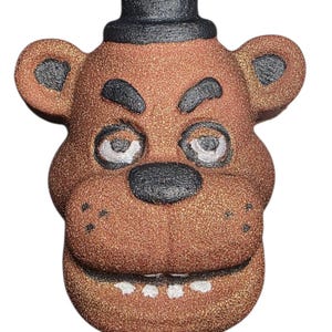 May include: A brown, glittery bear head with a black top hat, eyebrows, nose, and ear details. The bear has white eyes and a wide, toothy grin. This decorative item is inspired by a popular video game character.