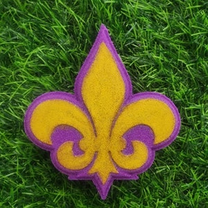 May include: A purple and yellow fleur-de-lis design. The design is made of a textured material, possibly sand.