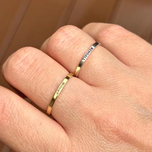 May include: Two thin gold rings are worn on a finger. One ring is engraved with the name "Willow", and the other with "Kendall". The rings are simple bands, likely made of gold or a gold-colored metal.