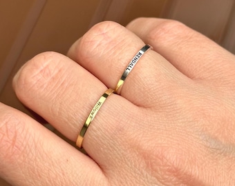 May include: Two thin gold rings are worn on a finger. One ring is engraved with the name "Willow", and the other with "Kendall". The rings are simple bands, likely made of gold or a gold-colored metal.
