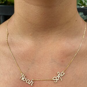 May include: A gold-toned necklace featuring two separate name pendants, Noah and Sofia, written in a cursive script. Each letter is encrusted with small, clear, sparkling stones. The pendants are connected by a delicate, thin gold-toned chain.