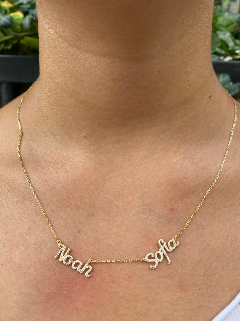 May include: A gold-toned necklace featuring two separate name pendants, Noah and Sofia, written in a cursive script. Each letter is encrusted with small, clear, sparkling stones. The pendants are connected by a delicate, thin gold-toned chain.