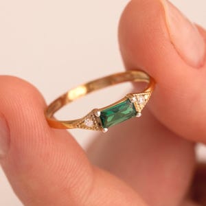 May include: A gold ring with a green emerald gemstone and small clear diamonds. The ring is on a finger.