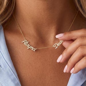 May include: A gold-toned necklace featuring two separate name pendants in a cursive script font, encrusted with small clear stones. The names Alayna and Deen are displayed on a delicate gold chain that rests against the neck.