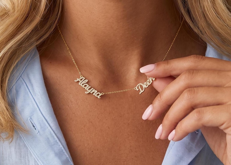 May include: A gold-toned necklace featuring two separate name pendants in a cursive script font, encrusted with small clear stones. The names Alayna and Deen are displayed on a delicate gold chain that rests against the neck.