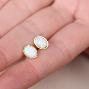 May include: A pair of oval-shaped opal earrings with gold settings. The earrings are shown on a person's finger.
