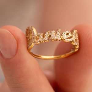 Pode incluir: Um anel de ouro com o nome "Marie" escrito em letra cursiva. As letras são incrustadas com pequenas pedras preciosas brilhantes. O anel é segurado entre dois dedos contra um fundo desfocado.
