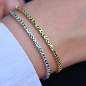 Pode incluir: Duas pulseiras de ténis, uma prateada e outra dourada, com pedras preciosas verdes. As pulseiras estão no pulso de uma pessoa.