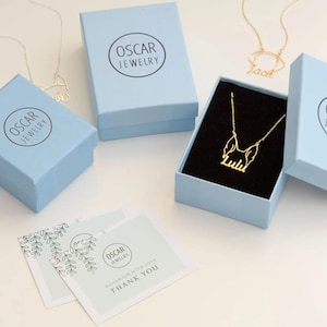 Peut inclure: Bo&icirc;tes &agrave; bijoux bleu clair avec le logo "Oscar Jewelry". L'une des bo&icirc;tes est ouverte, r&eacute;v&eacute;lant un collier en or avec le nom "Lulu". D'autres colliers sont visibles. Des cartes de remerciement sont &eacute;galement pr&eacute;sentes.