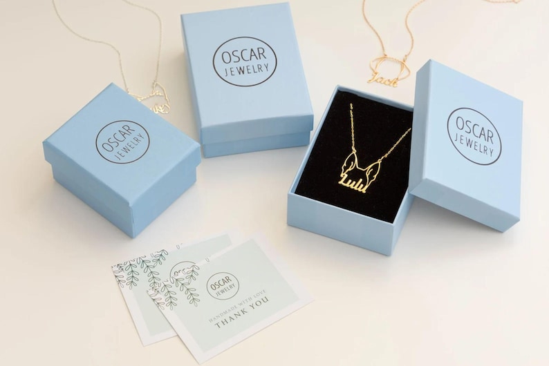Peut inclure: Bo&icirc;tes &agrave; bijoux bleu clair avec le logo "Oscar Jewelry". L'une des bo&icirc;tes est ouverte, r&eacute;v&eacute;lant un collier en or avec le nom "Lulu". D'autres colliers sont visibles. Des cartes de remerciement sont &eacute;galement pr&eacute;sentes.