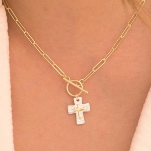 May include: A gold chain necklace with a toggle clasp and a silver cross pendant. The cross has a textured surface and a gold accent in the center.
