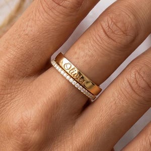 Pode incluir: Um conjunto de dois anéis dourados usados juntos. Um anel é de metal dourado liso e polido com o nome Olivia gravado em letra cursiva. O segundo anel é uma banda fina de estilo pavé cravejada com pequenas pedras transparentes.
