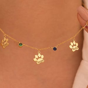 Puede incluir: Collar de oro con dijes de huella de pata y piedras preciosas de colores. Cada dije está grabado con un nombre: Kiko, Barney y Bella.