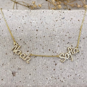 May include: A gold-toned necklace featuring two cursive name pendants, Noah and Sofia, encrusted with small clear stones. The names are connected by a delicate gold-toned chain, creating a horizontal design with a polished metallic finish.