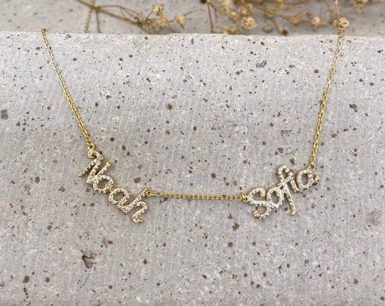 May include: A gold-toned necklace featuring two cursive name pendants, Noah and Sofia, encrusted with small clear stones. The names are connected by a delicate gold-toned chain, creating a horizontal design with a polished metallic finish.