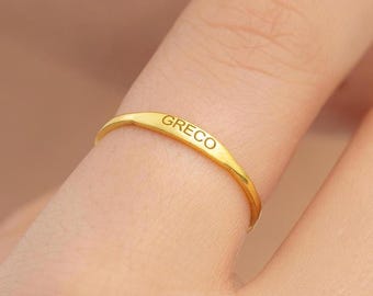 May include: A gold ring with the engraved word "GRECO" is worn on a finger. The ring is a thin band with a slightly curved design. The ring is a bright, shiny gold color.