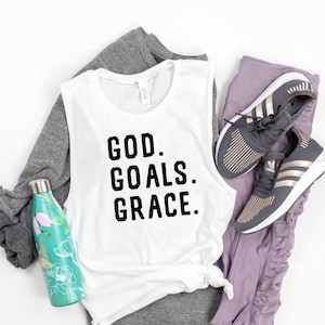 May include: White sleeveless tank top with the text "GOD. GOALS. GRACE." in black lettering. The tank top is paired with grey and purple athletic wear and a water bottle.