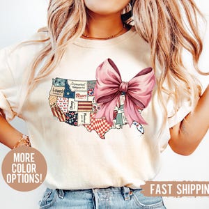 May include: A white t-shirt with a patchwork design of the United States map. The map is decorated with red, white, and blue patriotic patterns and a large pink bow. The text "Loved", "Empowered", "Brave", "Blessed", "Fierce", and "Precious" are visible on the map.