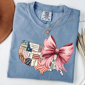 May include: A light blue t-shirt with a patchwork design of the United States featuring words like "Loved", "Brave", and "Empowered". A large pink bow is centered over the map.