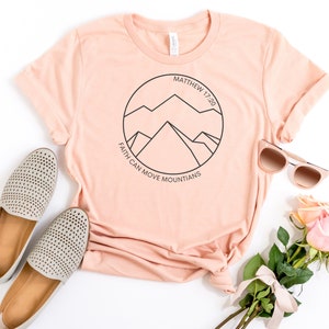 May include: A peach-colored t-shirt with a black circle graphic featuring a mountain range and the text "Matthew 17:20" and "Faith can move mountains".
