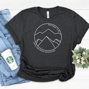 May include: A dark gray t-shirt with a white graphic of mountains inside a circle. The text "Matthew 17:20" and "Faith can move mountains" are printed around the circle.