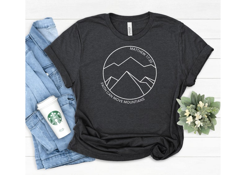 May include: A dark gray t-shirt with a white graphic of mountains inside a circle. The text "Matthew 17:20" and "Faith can move mountains" are printed around the circle.