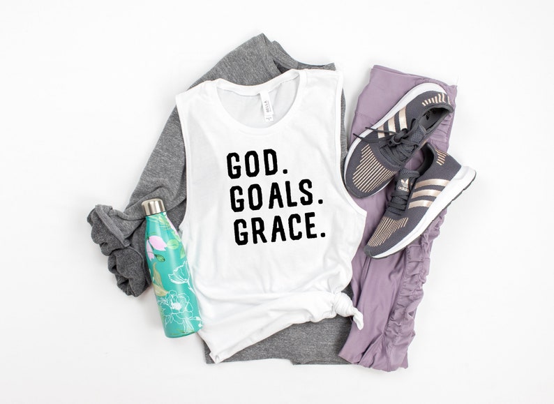 May include: White sleeveless tank top with the text "GOD. GOALS. GRACE." in black lettering. The tank top is paired with grey and purple athletic wear and a water bottle.