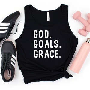 May include: Black tank top with the text "GOD. GOALS. GRACE." in white letters. The tank top is lying on a white surface with a pair of black trainers, a pink water bottle, and pink dumbbells.