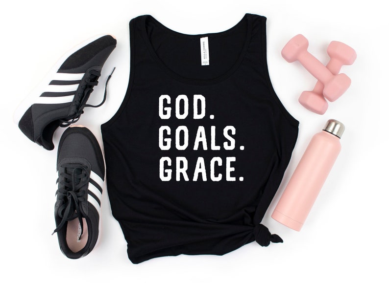 May include: Black tank top with the text "GOD. GOALS. GRACE." in white letters. The tank top is lying on a white surface with a pair of black trainers, a pink water bottle, and pink dumbbells.