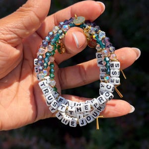 May include: A stack of three beaded bracelets featuring faceted crystal beads in shades of purple, teal, and clear. The bracelets include white square letter beads spelling out the phrases TRUST THE JOURNEY and THE PROCESS, with small gold-toned charms.