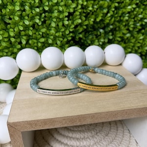 May include: Two stretch bracelets featuring light green beads and a central metal bar. One bar is silver-toned and the other is gold-toned. Both bars are engraved with the text ALL THINGS ARE WORKING FOR YOUR GOOD Rom 8:28.