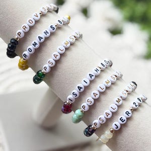 May include: A collection of seven stretch bracelets featuring white round beads with black printed text including PROV 3:5, ROMANS 8:28, PSALMS 34:18, ISAIAH 60:1, PSALMS 56:3, PSALMS 46:1, and JER 29:11, accented with small faceted colorful glass beads.