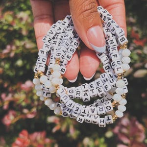 May include: A stack of four beaded bracelets featuring white square letter beads spelling out BLESSED, THANKFUL, and GRATEFUL. The bracelets are interspersed with small, round, translucent white faceted beads and tiny metallic gold spacer beads.