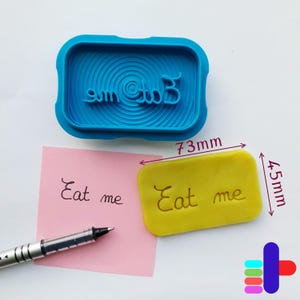 May include: A blue rectangular cookie stamp with the words "Eat me" in reverse. A yellow rectangular cookie with the words "Eat me" is next to a pink sticky note with the same text. The cookie is 7.3cm long and 4.5cm wide.