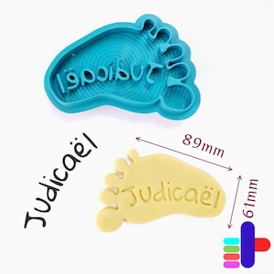 May include: A blue plastic foot-shaped cookie cutter with the name "Judicaël" in reverse. A yellow cookie with the same name and shape is shown, measuring 8.9cm long and 6.1cm wide. The name "Judicaël" is also written in black.