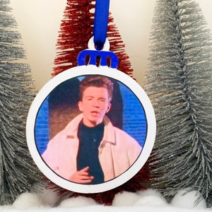 May include: A white round Christmas ornament with a blue ribbon and a photo of a man singing. The man is wearing a white jacket, a black turtleneck sweater, and has short brown hair.