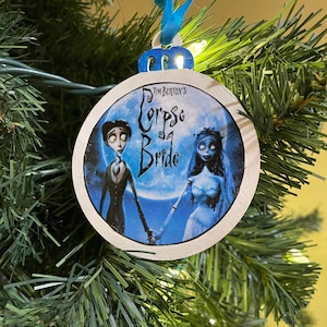 May include: A white round Christmas ornament with a blue ribbon. The ornament features a scene from the movie "Corpse Bride" by Tim Burton. The scene depicts the main characters, Victor and Emily, holding hands. The text "Tim Burton's Corpse Bride" is printed on the ornament.
