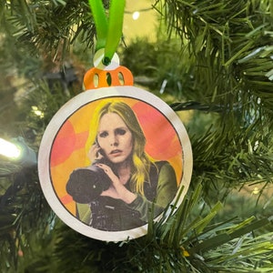 May include: A round Christmas ornament with a white background and a red, orange, and yellow design. The ornament features a woman with blonde hair holding a vintage camera.