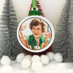 May include: A round white Christmas ornament with a picture of a woman smiling and holding rolling pins. The ornament has a green ribbon and a green hanger.