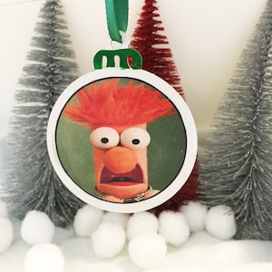 May include: A round Christmas ornament featuring a close-up image of the red-haired Muppet character, Beaker, with a surprised expression. The ornament is white with a green ribbon.