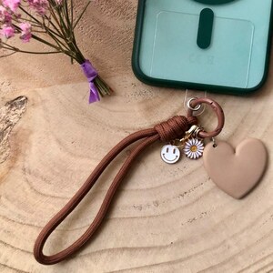 Peut inclure: Une lanière de téléphone marron avec un charm en forme de cœur, un charm en forme de marguerite et un charm en forme de smiley. Une coque de téléphone verte avec le texte "Soyez HAPPY & GIRL dans la vie!" est visible. Un petit bouquet de fleurs roses est également présent.